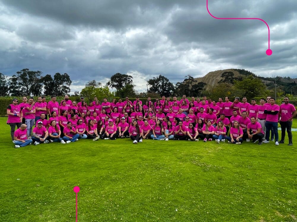Un gran grupo de personas con camisetas rosas posan para una foto en un campo de hierba con árboles y un cielo nublado de fondo.